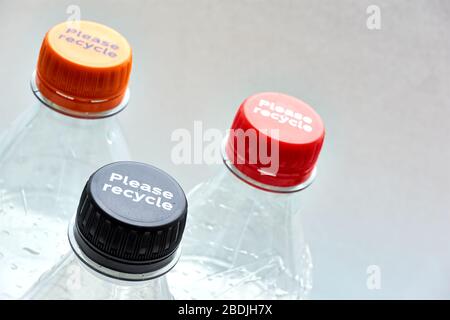 Plastic bottles and black, orange and red bottle caps with please recycle message against a grey background with copy space. Stock Photo