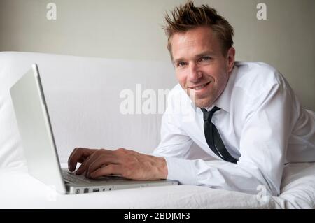 Friendly man reclining at home on comfortable white couch smiling with his laptop computer Stock Photo