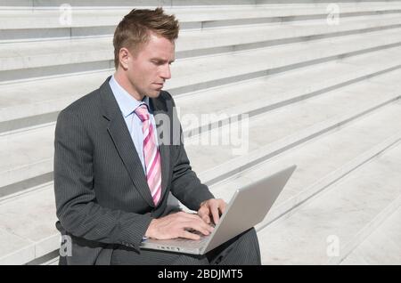 Serious businessman telecommuting outdoors sitting on sunny steps using his laptop computer Stock Photo