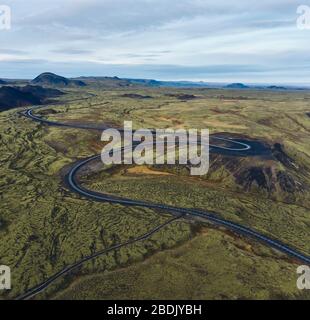 Drone view of narrow winding country road going through green mountainous terrain in Iceland with blue cloudy sky in background Stock Photo