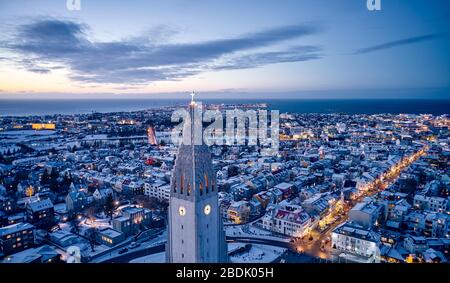 Drone view of white cathedral tower with cross above illuminated city of Reykjavik with ocean under endless sky Stock Photo