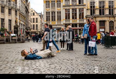 Brussels, Belgium - October 12 2019: Woman tourist taking photo of her friends via cellphone by lying on ground in grand place