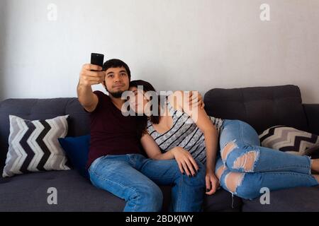 Young adult Hispanic married couple hugging on the couch watching Tv Stock Photo