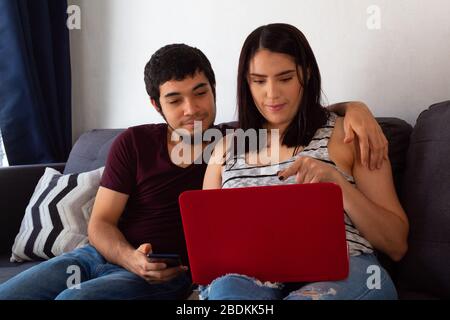 Young adult Hispanic married couple working from home together with a laptop on the sofa at home because of the pandemic Stock Photo