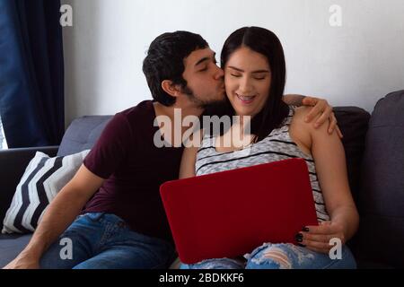 Young adult Hispanic married couple working from home together with a laptop on the sofa at home, kissing each other on the cheek Stock Photo