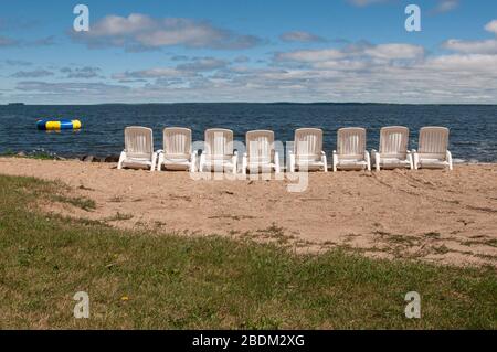 Eight empty plastic chairs lined up facing the water on a sandy beach with a colorful inflatable swimming float on the water in the distance. Stock Photo