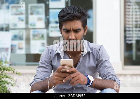 Man phone. Closeup portrait angry indian male holding texting looking at cellphone isolated outdoors background Stock Photo
