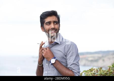 Happy ethnic man smoking cigarette, smiling, thinking looking sidewards outdoors background. Stock Photo