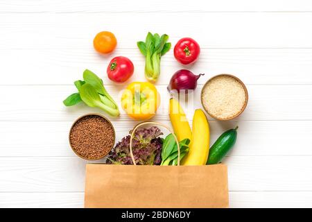 Full eco paper bag of different health food - buckwheat, rice, yellow bell pepper, tomatoes, bananas, lettuce, green, cucumber, onions on white wooden Stock Photo