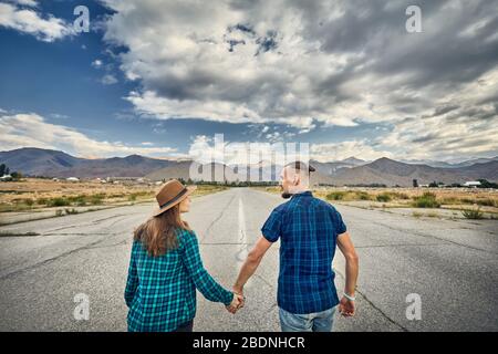 Happy couple in checked shirt holding by hands and walking on the wide asphalt road with mountains and cloudy sky background Stock Photo