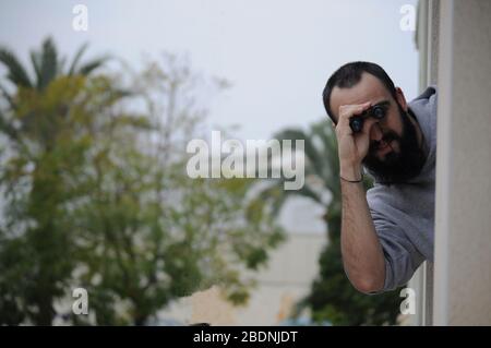 bald man with long beard leaning out from building window looking through binoculars. Picture with copy space of man using binoculars to spend time on Stock Photo