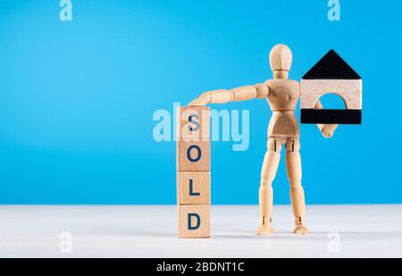 Wooden man hold mini house. Mannequin put his hand on tower of cubes with inscription SOLD. Concept of property, earnings, rent, business, mortgage an Stock Photo