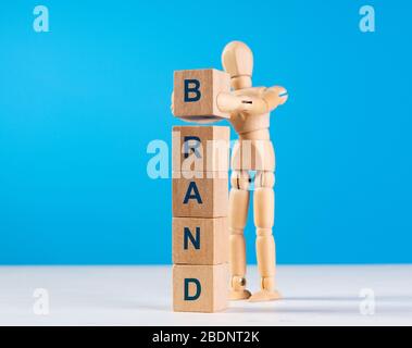 Wooden man construct tower with blue word BRAND. Mannequin put last block on a top. Concept of brand building for success, brand awareness improvement Stock Photo