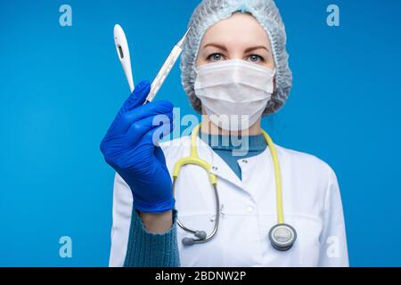 Beautiful woman doctor in white medical clothes, mask, phonendoscope and medical hat shows electric thermometers, picture isolated on blue background Stock Photo