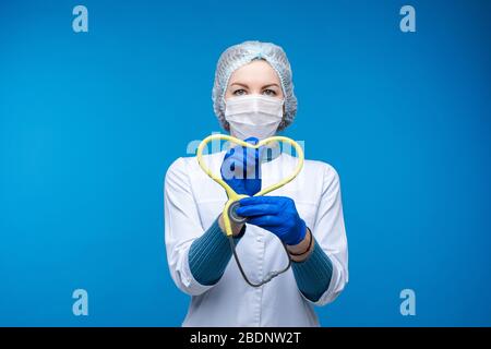 Beautiful woman doctor in white medical clothes, mask holds a phonendoscope and shows a heart, picture isolated on blue background Stock Photo