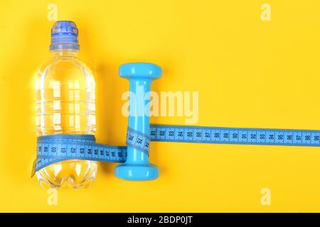 Sports tools in cyan blue. Bottle of water and fitness equipment on yellow background. Shaping and refreshment concept. Centimeter wrapped around bottle and dumbbell, top view. Stock Photo