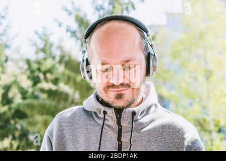 Bearded man in hoodie with headphones listening to music in park outdoors, head and shoulders Stock Photo