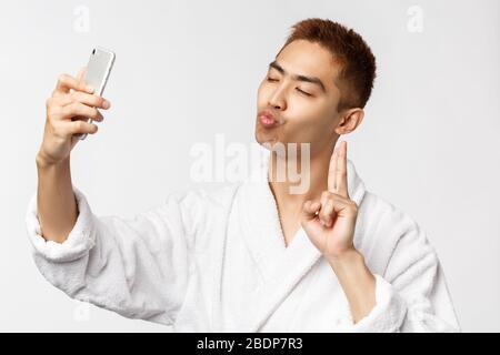 Beauty, spa and technology concept. Cheeky and silly asian man in bathrobe, close eyes and making kissing face taking selfie, show peace sign at Stock Photo
