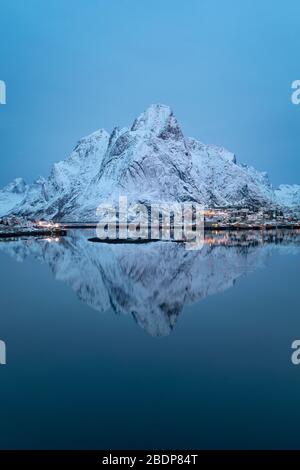 Lonely mountain perfectly reflected in cold, calm waters of northern fjord. Snow covered mountain ridge over early morning calm bay of Reine, Norway Stock Photo