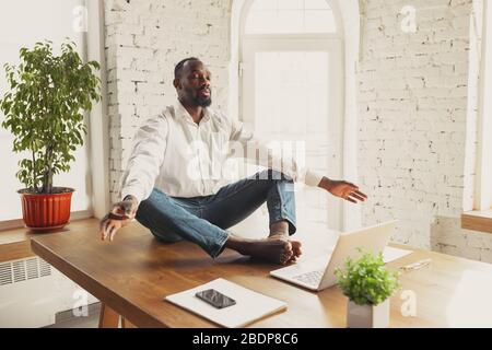 Young african-american man doing yoga at home while being quarantine and freelance online working. Remote, isolated or alone at office. Concept of healthy lifestyle, wellness, activity, movement. Stock Photo