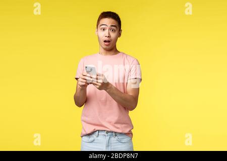 Technology, communication and lifestyle concept. Surprised asian male in pink t-shirt, holding mobile phone, open mouth gasping wondered, react to Stock Photo