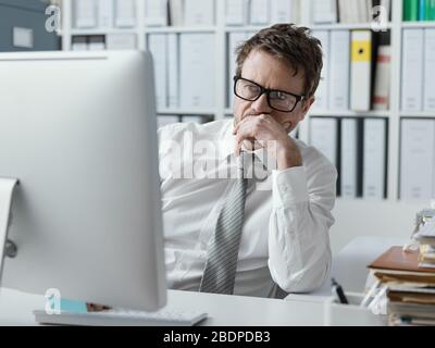 Pensive businessman working in the office and staring at the computer screen with hand on chin Stock Photo