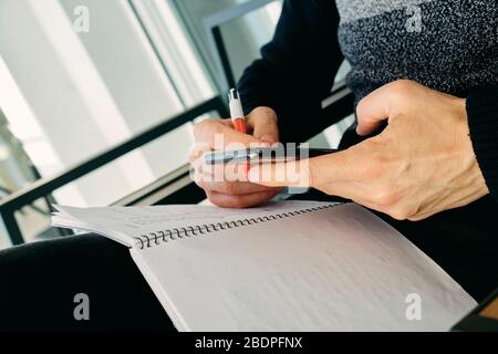 closeup of a young caucasian man, in casual wear, using his smartphone while is taking notes in a notebook, sitting in an outdoors armchair on the bal Stock Photo