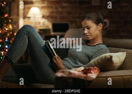 Woman lying on the sofa and reading a book she received as Christmas gift, she has just unwrapped it Stock Photo