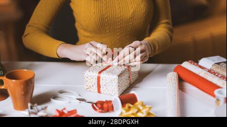 Woman wrapping Christmas gifts at home, she is tying a ribbon bow: holidays and celebrations concept Stock Photo