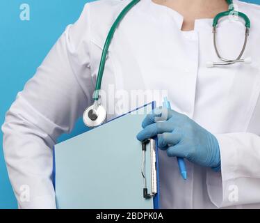 medic woman in white coat, blue latex gloves holding a tablet with a clip for papers, blue background, close up Stock Photo