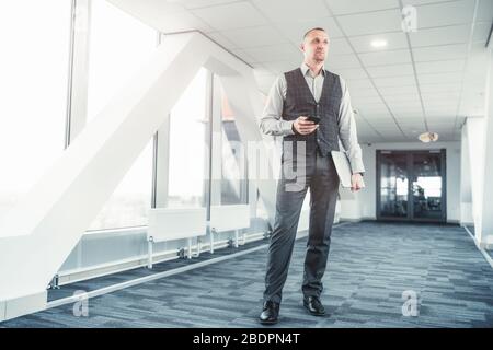 A tall handsome elegant man caucasian entrepreneur is standing in a bright passageway with a smartphone and laptop in his hands Stock Photo