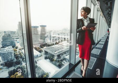 A good-looking African-American woman entrepreneur in a red skirt and black jacket is using a digital tablet while leaning against a panoramic window Stock Photo