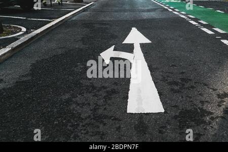 Road marking on the asphalt: white paint branch arrow pointing forward and to the left, a fragment of a green bicycle lane on the right Stock Photo