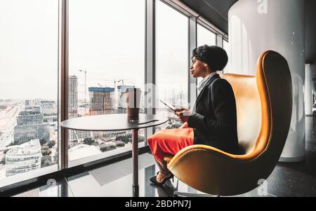 A ravishing African-American businesswoman on a yellow armchair is using her tablet PC; biracial woman entrepreneur using a digital tablet Stock Photo