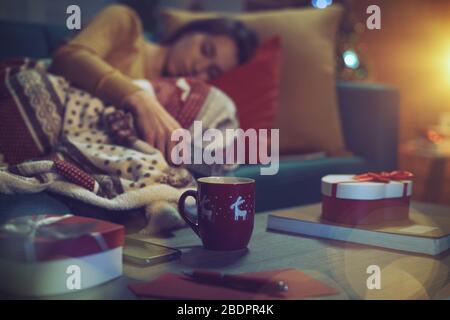 Young woman lying down on the sofa and falling asleep on Christmas Eve, she is holding the TV remote control, gifts in the foreground Stock Photo