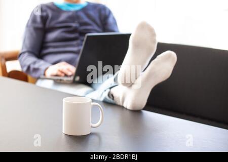 Woman sitting in a couch with a laptop computer an her feet up on a table where a mug or cup is placed. Selective focus and woman blurred. Working fro Stock Photo