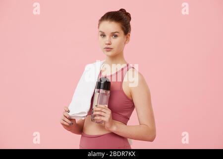 Portrait of fit young woman with towel and bottle of water looking at camera over pink background Stock Photo