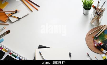 Top view image of painter working desk. Color tube, Potted plant, Paint brush, Stationary, Color palette and Notebook flat lay on white isolated backg Stock Photo