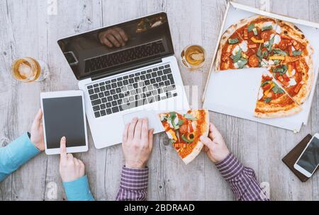 Office workers having a lunch break in the office, they are eating pizza, using a laptop and a digital tablet, flat lay Stock Photo