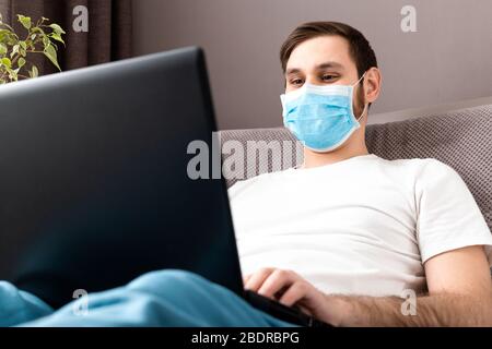 Young caucasian man working from home office wearing protective mask using laptop and internet. Coronavirus covid 19 quarantine. Remote work Stock Photo