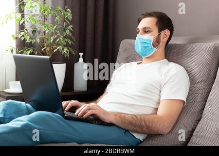 Young caucasian man working from home wearing protective mask using laptop and internet. Cozy Home office, workplace on sofa during coronavirus Stock Photo