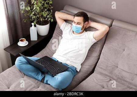 Happy relaxed young caucasian man working from home wearing protective mask using laptop and internet. Cozy Home office, workplace on sofa during Stock Photo