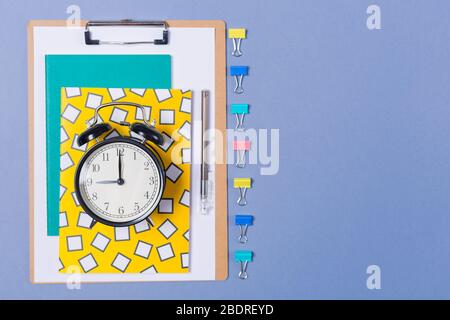 Pile of bright notebooks, clipboard, alarm- clock and stationary on blue desk top. Copy space. Free space. Start of working day.  Stock Photo