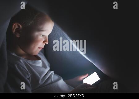 A child under a blanket playing with a smartphone, sitting on the Internet. The concept of spending time in safe isolation. Stock Photo