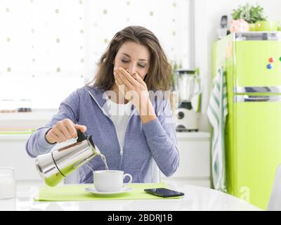 Tired lazy woman having breakfast at home in the kitchen, she is yawning and pouring coffee Stock Photo