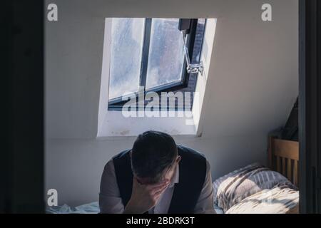 A man looking down with head in hand, wearing a waistcoat sitting on a bed with a window behind. Mental health concept. Stock Photo