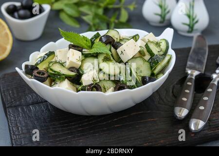 Healthy salad with cucumbers, feta cheese and black olives, with olive oil and greens, located in white bowl on a dark background, horizontal Stock Photo