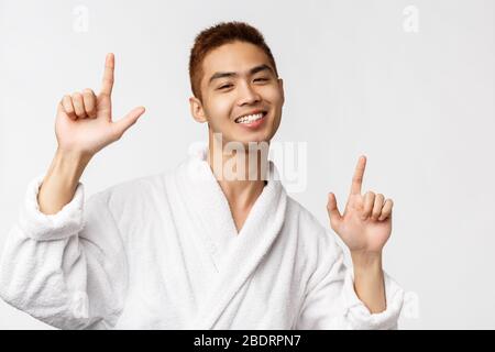 Beauty, spa and leisure concept. Portrait of cheerful, handsome enthusiastic asian man in bathrobe, pointing fingers up, showing way and dancing Stock Photo