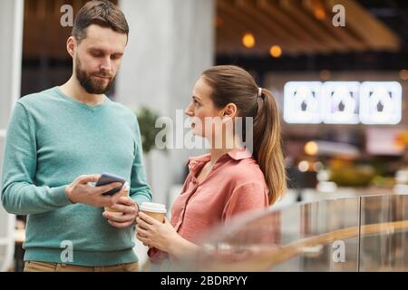 Young woman drinking coffee and talking to man while he using his mobile phone they are in the mall Stock Photo