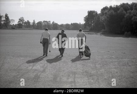 1960s, historical, on a golf course, in a line, three men walking down a fairway, two golfers and a caddie who is carrying clubs for one of them  and who is wearing a suit, while the other golfer is pulling a golf bag on a trolley of the era. Stock Photo